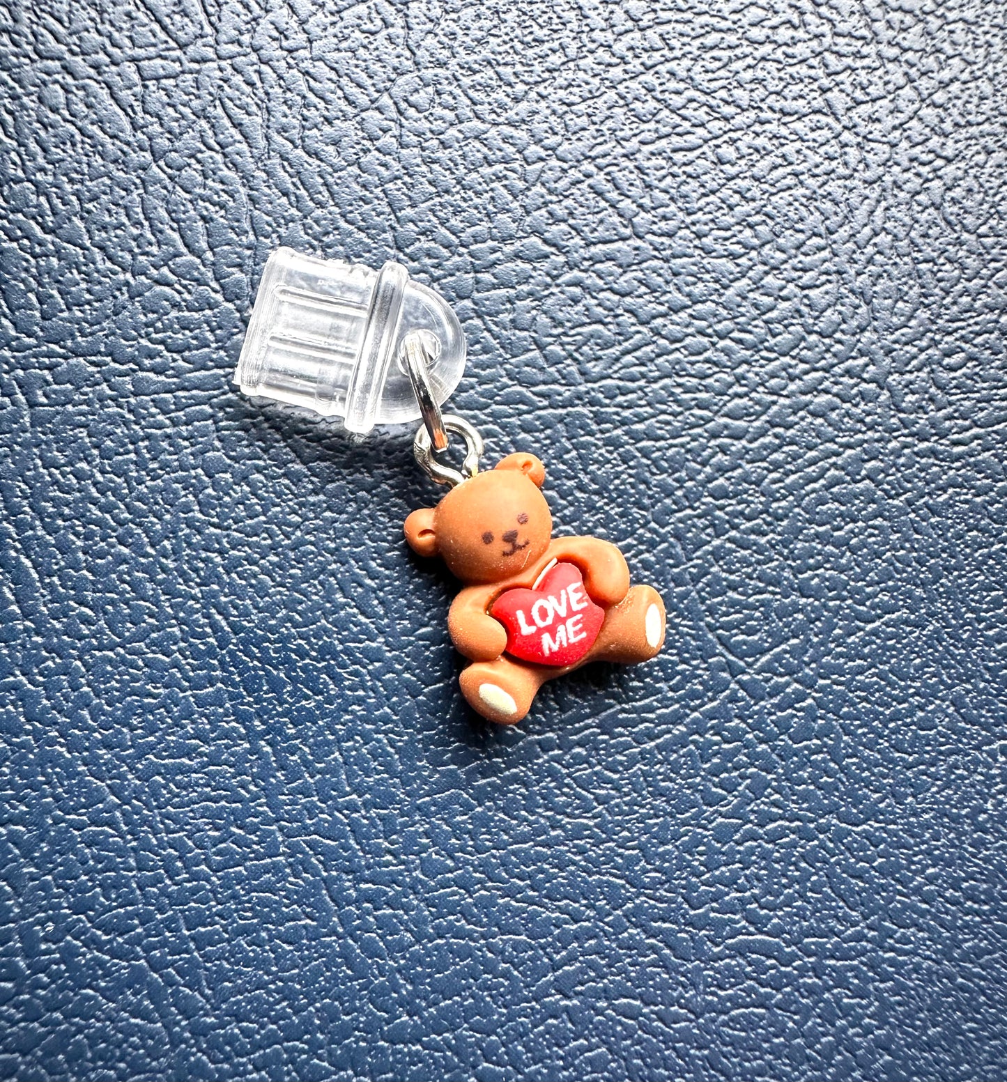 Small teddy bear charm with a heart that says 'LOVE ME' on a blue textured background