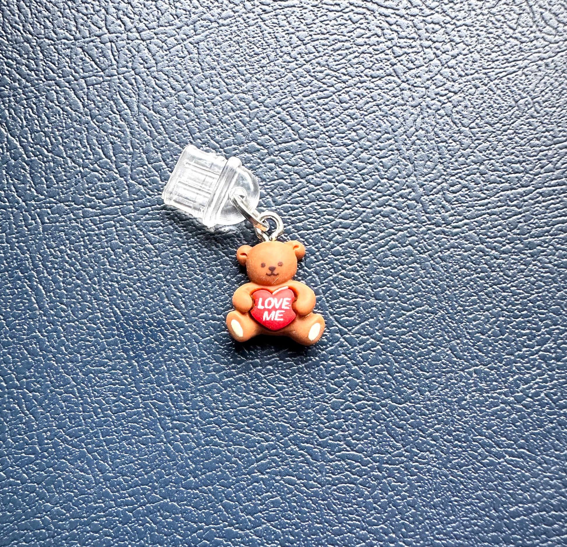 Small teddy bear charm with a heart that says 'Love Me' on a textured blue background